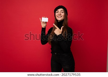 Photo of beautiful young brunette woman wearing black sweater and hat isolated on red background holding credit card looking at camera and pointing finger at plastic card