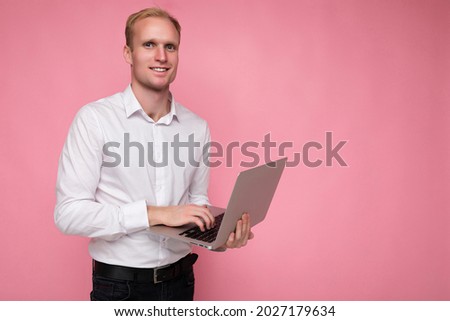 Side profile photo shot of handsome smiling confident blonde adult male person holding computer laptop typing on keyboard wearing white shirt looking at camera isolated over pink background