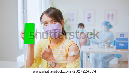 Immunity vaccine concept - Happy Asian young women sitting smile showing adhesive plaster her arm and thumbs up gesture after vaccinated for covid19 and smartphone displaying green screen in hospital