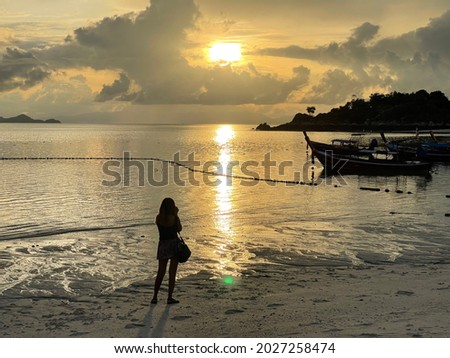 A woman stands to take pictures of the sunrise by the sea and the long tail boat.