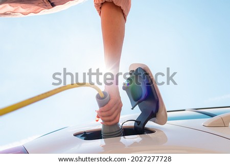 Low angle shot of a Caucasian woman opening an electric car charging socket cap and plugging in a charger Royalty-Free Stock Photo #2027277728
