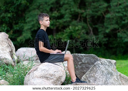 Boy sitting on a stone with laptop in garden. Kid have online web lesson or class on computer at nature, green class. Back to school. Outdoor learning concept