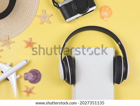Top view of white cover book  with headphones around, camera, straw hat, airplane model sea shells and starfishes on yellow background. Traveling audio book or podcast background.