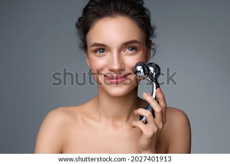 Attractive woman massaging her cheek with massage Y-shaped ball roller. Photo of woman with perfect skin on gray background. Beauty and skin care concept