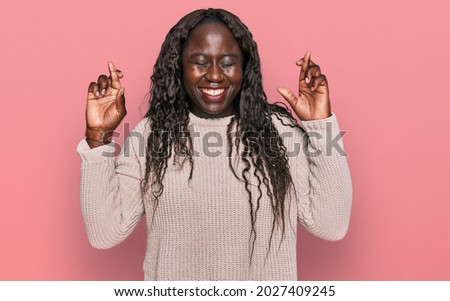 Young african woman wearing wool winter sweater gesturing finger crossed smiling with hope and eyes closed. luck and superstitious concept. 