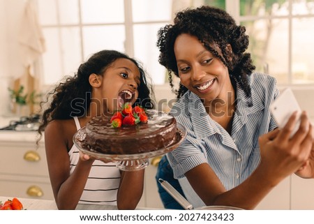 Beautiful mother with cute girl taking selfie holding a chocolate cake in the kitchen. Happy young family of mother and daughter taking self portrait while baking cake.