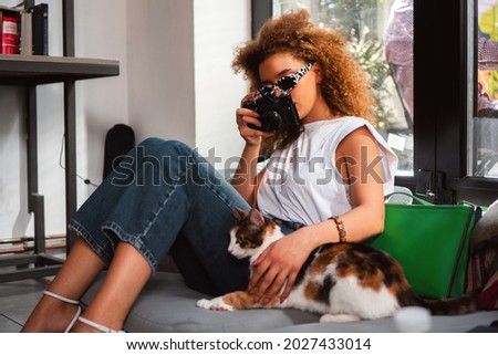 A young woman sits on the sofa and takes photos of her cat with a photo camera.