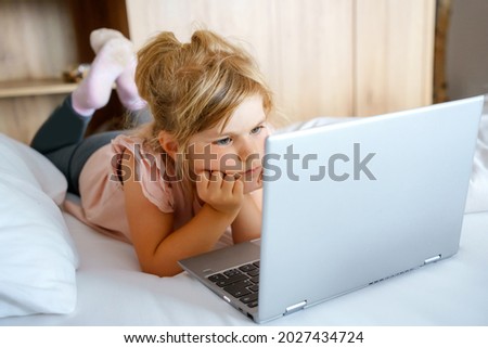 Preschool girl watching videos on laptop, notebook,in bed on clean white linens. Indoors activity with children. Freelance, distance learning or work from home with kids concept. Happy child