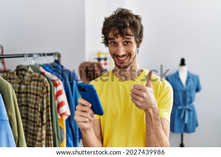 Young hispanic man at retail shop using smartphone smiling happy and positive, thumb up doing excellent and approval sign 