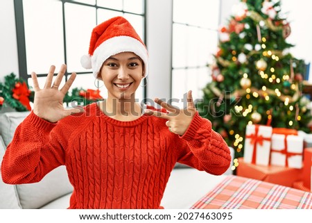 Young hispanic woman with short hair wearing christmas hat sitting on the sofa showing and pointing up with fingers number eight while smiling confident and happy. 