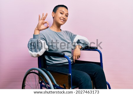 Beautiful hispanic woman with short hair sitting on wheelchair smiling positive doing ok sign with hand and fingers. successful expression. 