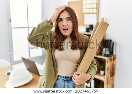 Young brunette woman holding paper blueprints at the office stressed and frustrated with hand on head, surprised and angry face 
