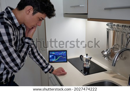 worried young man on phone while making coffee in kitchen and stock data on screen