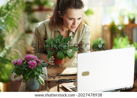 Relaxing home gardening. 40 years old woman in white rubber gloves with potted plant and laptop at modern home in sunny day.