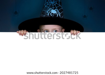 A girl wearing a witch hat peeps out from behind a white horizontal wall. Dark background and white copy space for your design. Halloween banner.
