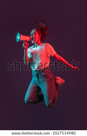 High jumps. Vertical portrait of beautiful girl in casual clothes jumping isolated on dark purple studio background in red neon light, filter. Concept of emotions, facial expression, beauty, sales.