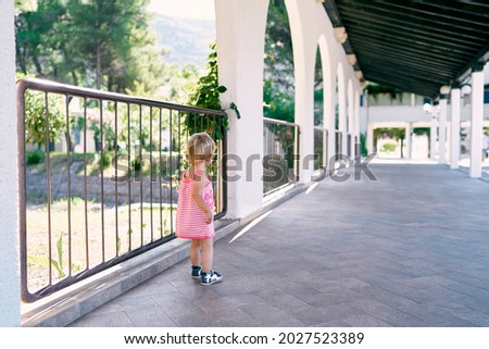 Little girl stands near a fence in a long pavilion in the park. Back view
