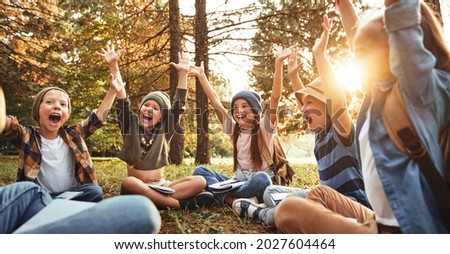 Excited school kids sitting on grass in forest with notebooks during outdoor science lesson, raising arms and screaming from excitement, discussing discoveries with teacher and learning ecology basics Royalty-Free Stock Photo #2027604464