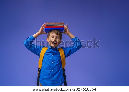 Back to school. A cheerful schoolboy against a purple paper wall. A child with a backpack, books and notebooks. The boy is ready to study.