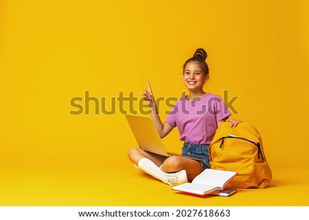 happy schoolgirl with backpack, laptop and books is sitting on yellow background in the Studio. The child shows, a sign of approval, a finger up. Back to school. copy space.