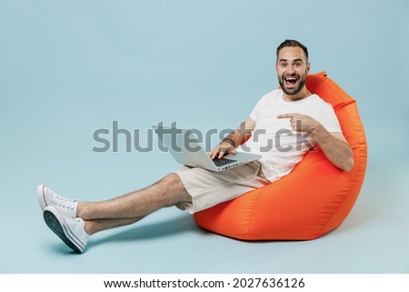 Full length young surprised happy man in casual white t-shirt sit in bag chair hold use work point index finger on laptop pc computer rest relax isolated on plain pastel light blue background studio.