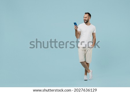 Full length young smiling friendly happy man 20s wearing casual white t-shirt looking camera using mobile cell phone chat onine isolated on plain pastel light blue color background studio portrait. Royalty-Free Stock Photo #2027636129