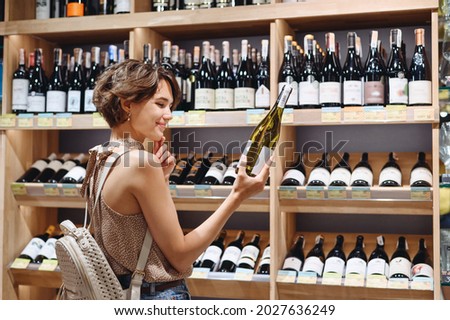 Smiling young woman wearing casual clothes shopping at supermaket grocery store buy choosing white wine alcohol hold bottle prop up chin inside hypermarket. People purchasing gastronomy food concept Royalty-Free Stock Photo #2027636249