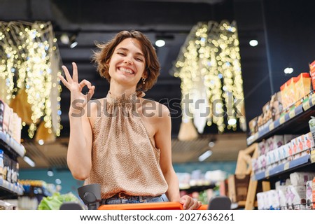 Young satisfied smiling happy fun european woman in casual clothes shopping at supermaket store with grocery cart show ok okay gesture inside hypermarket. People purchasing gastronomy food concept.