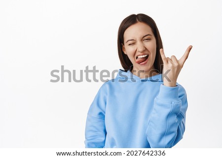 Cheerful and positive girl shows horns heavy metal sign, winking and showing tongue, having fun, enjoy event and celebrating, standing in hoodie against white background