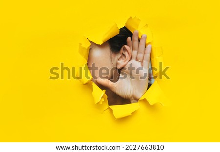 Close-up of a woman's ear and hand through a torn hole in the paper. Yellow background, copy space. The concept of eavesdropping, espionage, gossip and tabloids. Royalty-Free Stock Photo #2027663810