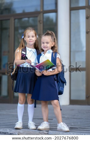 Two girls schoolgirls stand in front of the school building and look at a notebook Royalty-Free Stock Photo #2027676800