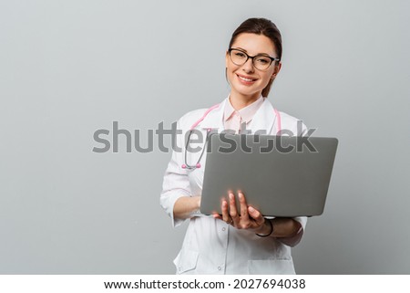 Portrait of a friendly smiling young female doctor. Image of a female doctor with glasses and with a laptop. Isolated on a grey background. Royalty-Free Stock Photo #2027694038