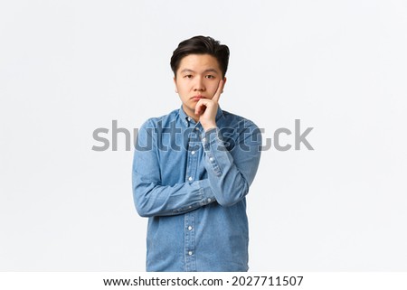 Bored and uninterested asian male in blue shirt, looking unamused and careless at camera, listening boring speech, standing displeased and annoyed over white background, tired of person
