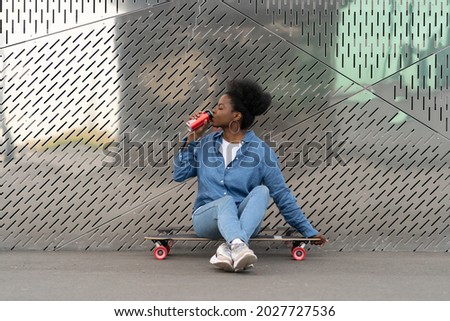 Tired african skateboarder girl refreshing after longboarding sit on skate drinking soda beverage from metal can. Trendy urban young female relaxing after active training on longboard in urban space
