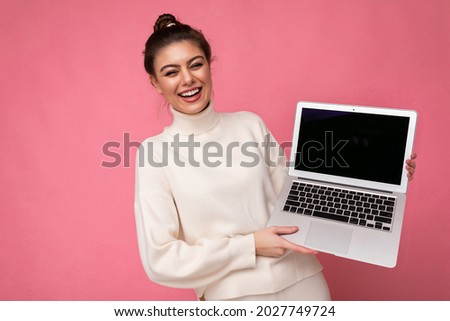 Photo of beautiful smiling and laughung brunette young lady with gathered dark hair wearing white sweater holding computer laptop and looking at camera with close eyes and open mouth isolated on pink