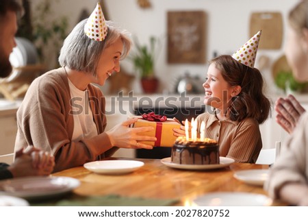 Cute little girl receiving congratulations from grandma at home, loving senior woman giving gift to excited happy granddaughter during Birthday celebration with family, cake with candles on table