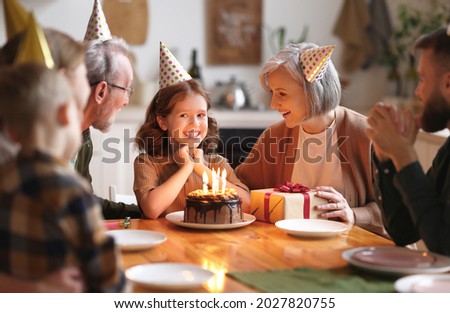Happy little girl celebrating birthday with family at home, looking at cake with lit candles, making wish while sitting at table in kitchen with smiling loving parents, grandparents and small brother