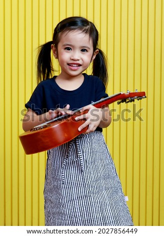 Portrait studio shot of little cute pigtails hairstyle preschooler musician happy girl standing smiling holding small dark brown wooden ukulele in hands posing in front yellow stripe wall background.