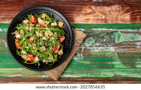 close up picture of fresh rocket salad with tomato, crouton, herb roasted chicken, and parmesan cheese in black ceramic bowl on sack cloth on colourful wooded table. concept for healthy.