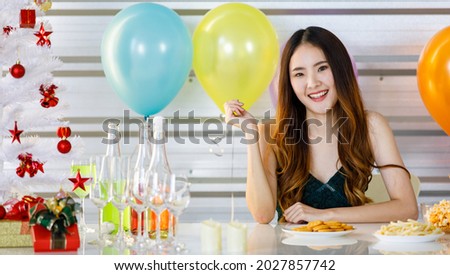 Asian beautiful girl sit smile rest chin on hand at beverage table with alcohol drinks bottles tall wine glasses cookies french fries gift in front colorful balloons and xmas tree at Christmas party.