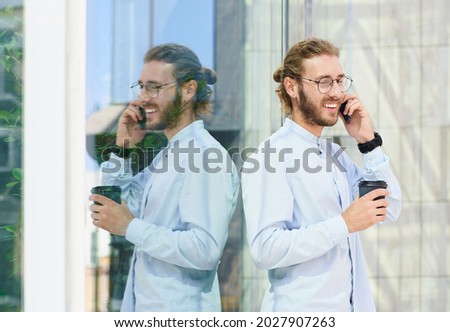 Smiling, in a good mood, a young businessman in a blue shirt, drinking coffee and talking on the phone, posing in the street, near a glass building. Business and people concept.