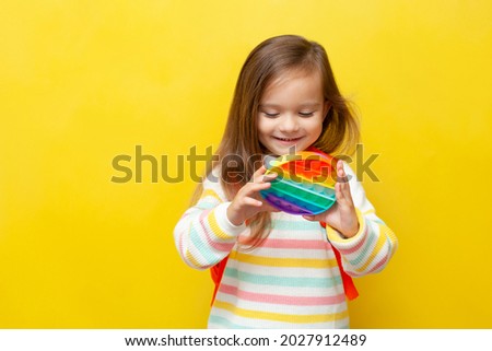 A little schoolgirl in a striped blouse and with a backpack holds a pop it in her hands and smiles. Anti-stress. Stress. Back to school. Brightly. Yellow background, space for text.