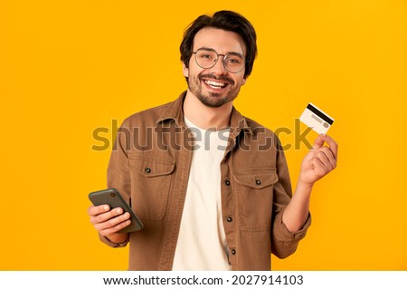 Young bearded man in glasses holds credit card and smartphone isolated on orange background. Online shopping. Online payment. Royalty-Free Stock Photo #2027914103