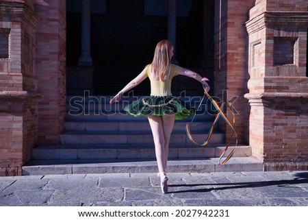 Hispanic adult female classical ballet dancer in green and black tutu with coins, dancing with a rhythmic gymnastics ribbon in the colors of the gay pride flag.