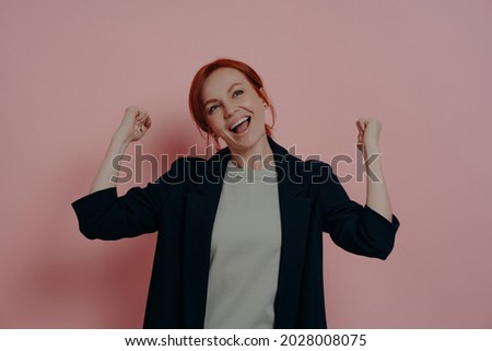 People and achievements. Indoor shot of young happy overjoyed ginger business woman raising hands in fists and celebrating triumph, screaming in excitement, isolated over pink studio background.