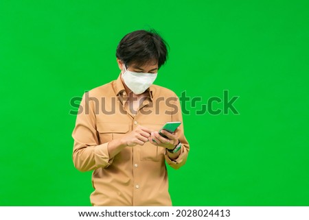 A half-body of an Asian man in a yellow shirt holding his phone wearing a medical mask stands behind a green screen.