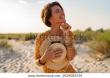 stylish attractive slim smiling woman on beach in summer style fashion trend outfit carefree and happy, feeling freedom, wearing yellow printed dress boho style chic and straw hat