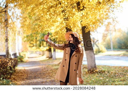 Fashionable woman is taking selfie while spending time outdoors in autumn, stylish girl is wearing wide brimmed hat and beige coat. 