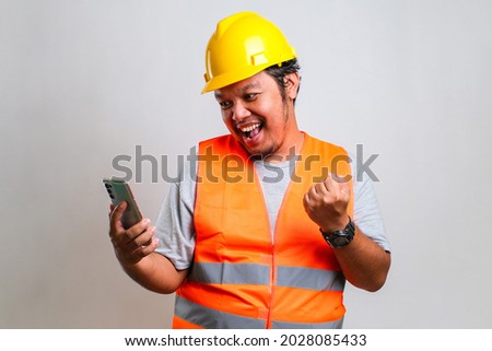 Young asian engineer worker wearing orange vest and helmet over isolated white background celebrating surprised while looking at his phone, amazed for success with left arm raised Royalty-Free Stock Photo #2028085433