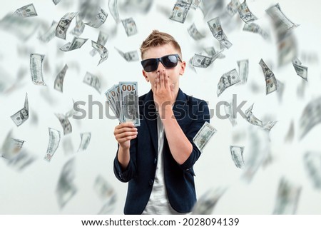 A cute, rich boy with glasses holds dollars and knows how to make money against the background of falling banknotes. Money rain, Financial education, young millionaire, investments, loans, winnings